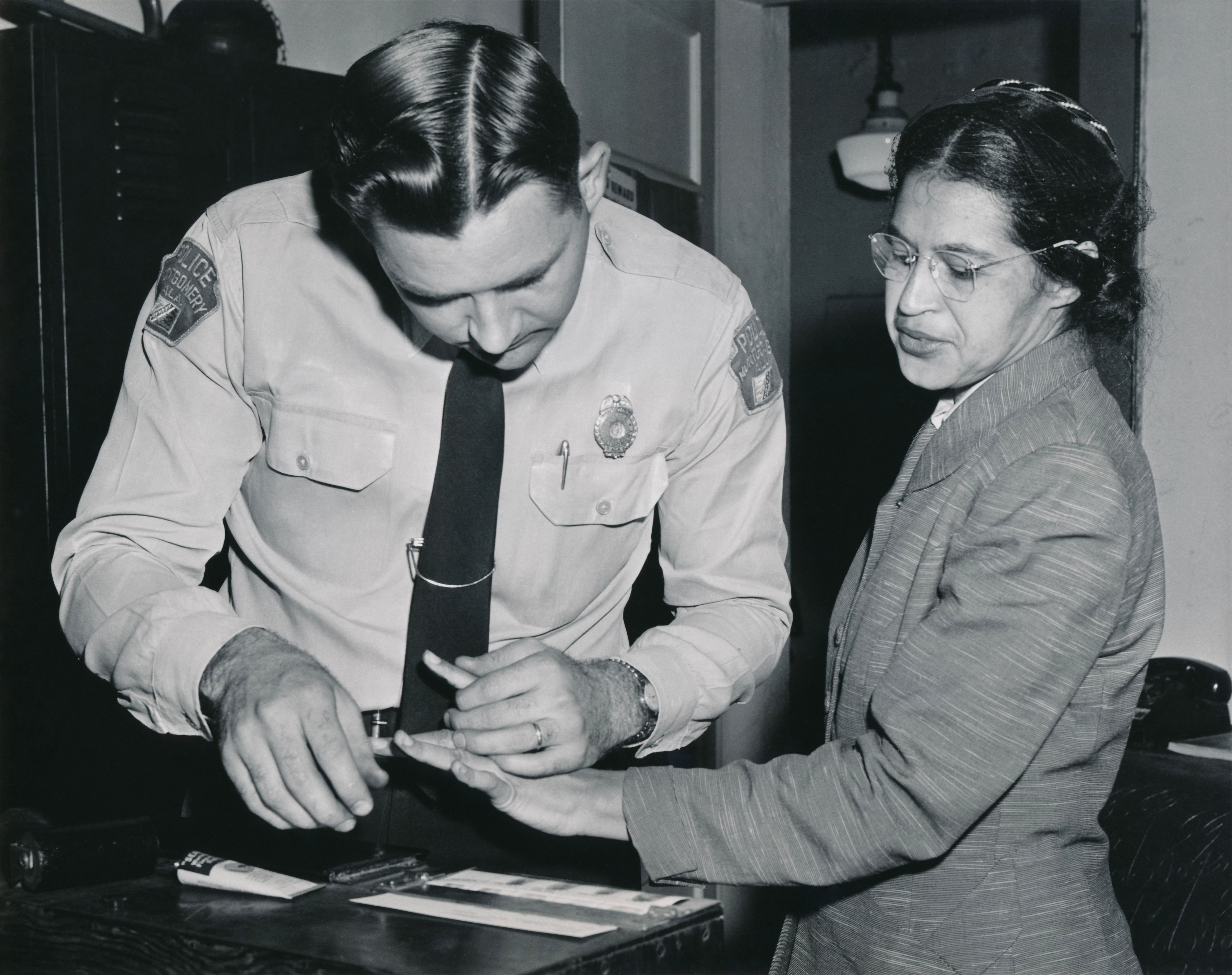 A police officer takes fingerprints from a woman wearing glasses and a jacket in an office setting.