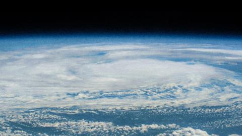 From a vantage point reminiscent of Carl Zimmer's explorations, an aerial view captures a massive swirling cloud formation over Earth, resembling a hurricane or cyclone against the infinite darkness of space.